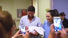 新手爸爸抱着刚出生的宝宝给产房外的人看，大家露出喜悦的表情