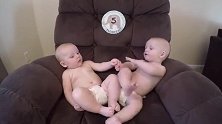 肉嘟嘟双胞胎坐沙发上，望着彼此牵手互撩啃脚丫，玩的不亦乐乎